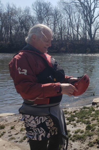 Photo of author with DuPage River in background. Author is 6 foot-0 inches, 230 pounds, long gray hair in a bun. He is wearing red dry top, red life jacket and gray splash jacket, and is preparing to don a red helmet.